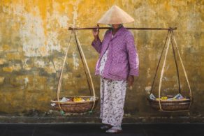 Habitante de Hội An portant un chapeau conique traditionnel vietnamien et un panier suspendu, scène de vie locale au cœur du Vietnam entre culture, artisanat et voyage sur mesure en Asie.