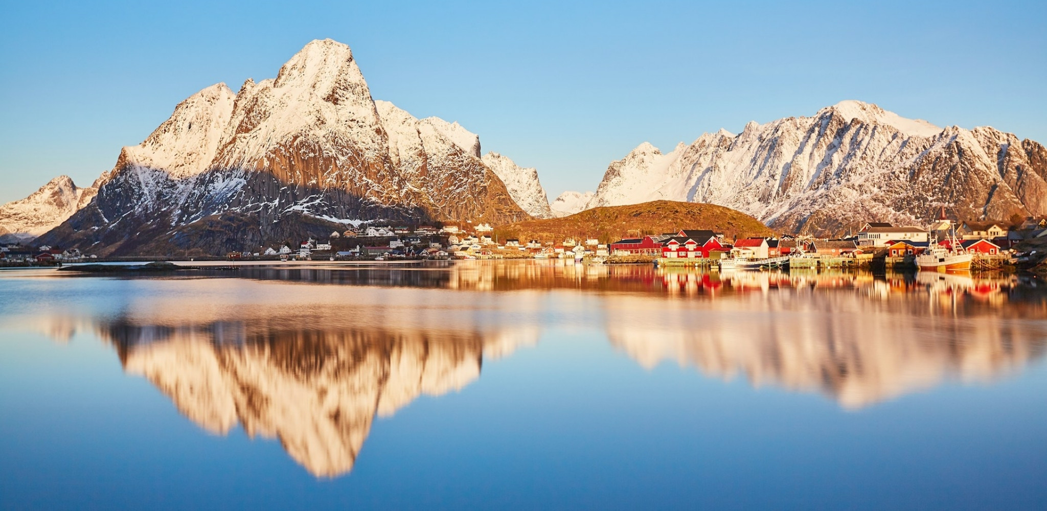 Village de pêcheurs des îles Lofoten au pied de montagnes enneigées se reflétant dans un fjord calme.