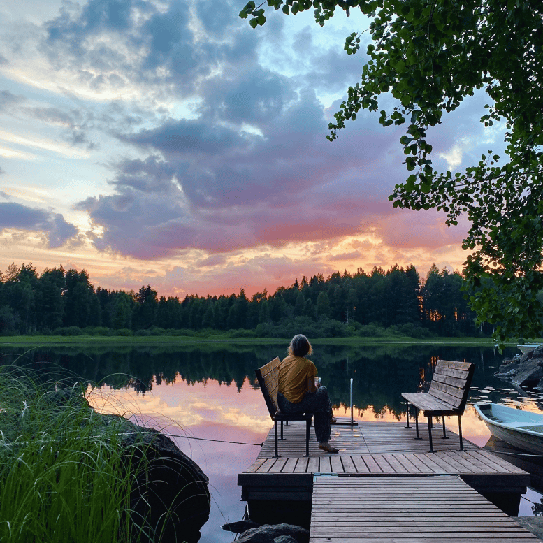 Personne contemplant le coucher du soleil depuis un ponton en bois au bord d'un lac paisible, entouré de forêts en Finlande.
