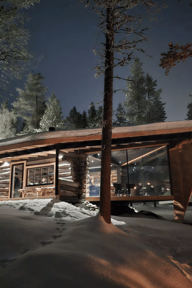 Chalet contemporain en bois aux grandes baies vitrées, illuminé à la tombée de la nuit au milieu d'une forêt enneigée en Finlande.