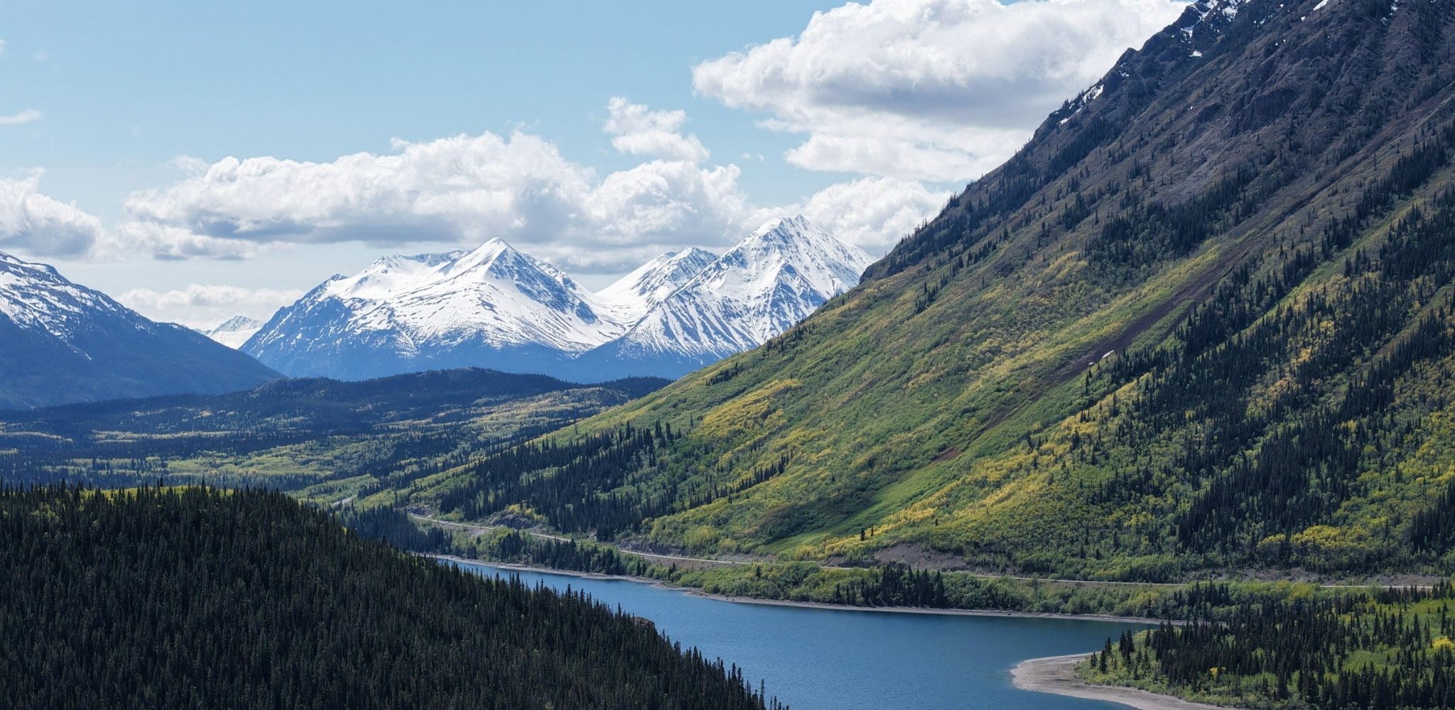 Paysage du Yukon composé d'une vallée traversée par une rivière, bordée de forêts boréales et de pentes verdoyantes, avec une chaîne de sommets enneigés se dressant à l'horizon sous un ciel parsemé de nuages.