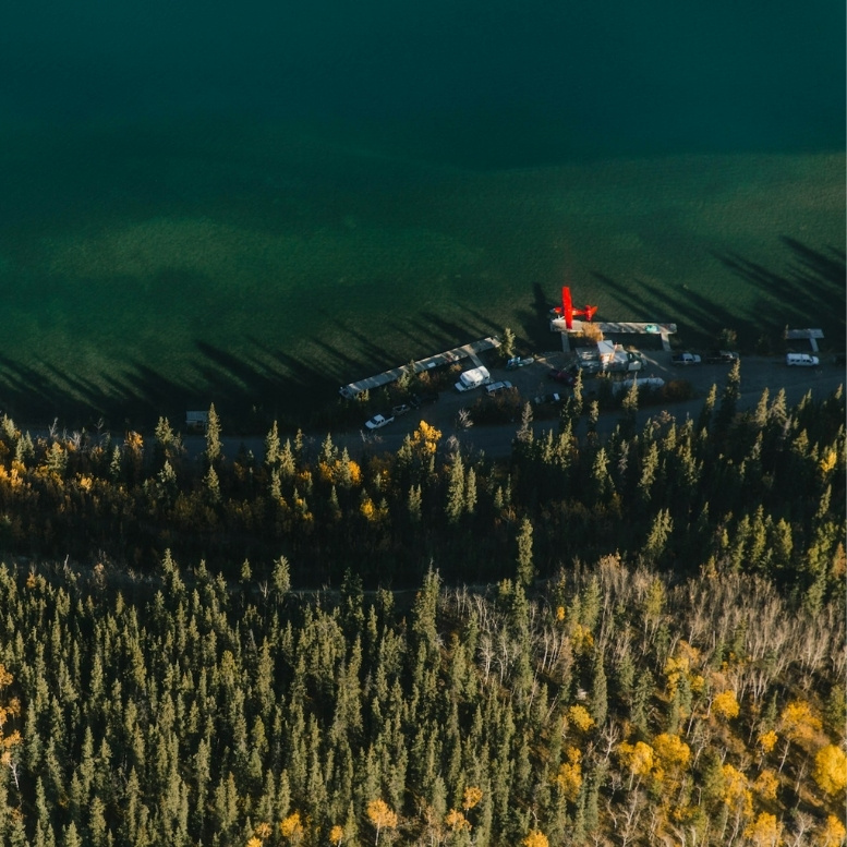 Vue aérienne d'une forêt boréale du Yukon aux couleurs d'automne, bordant une rivière aux eaux vert émeraude. Un hydravion rouge est amarré sur une petite base isolée, illustrant le caractère sauvage et reculé du nord canadien.