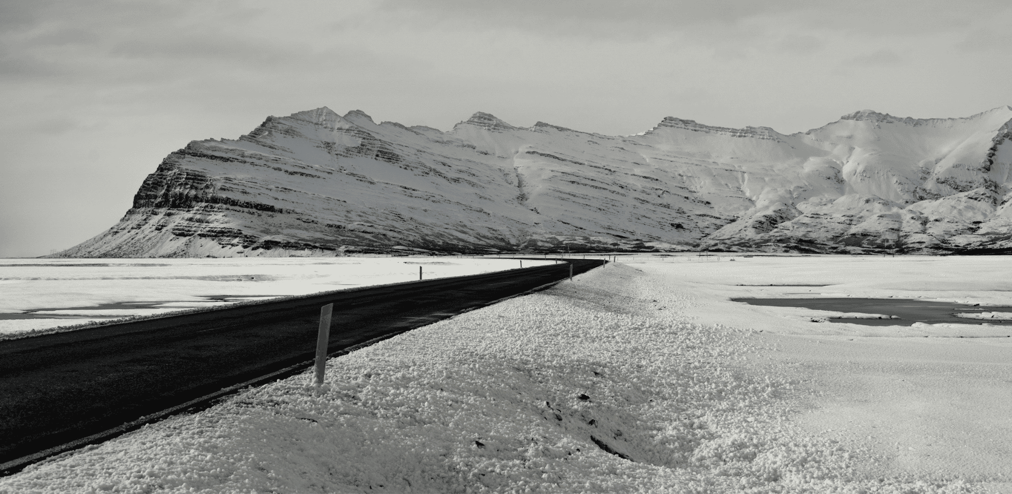 Route asphaltée traversant une vaste plaine enneigée en Islande, au pied d'une imposante chaîne de montagnes aux sommets recouverts de neige.