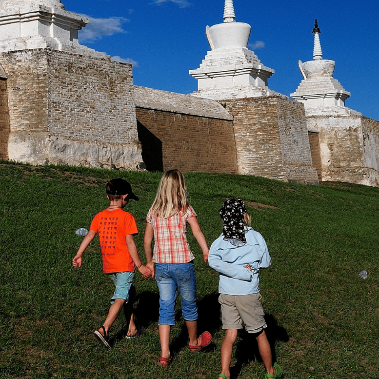 Trois enfants découvrant un monastère bouddhiste traditionnel en Mongolie, avec ses stupas blancs sous un ciel bleu.